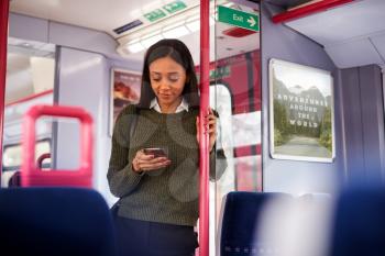 Female Passenger Standing By Doors In Train Looking At Mobile Phone