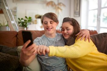 Young Downs Syndrome Couple Sitting On Sofa Using Mobile Phone To Take Selfie At Home