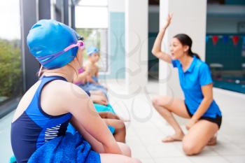 Female Coach Giving Children In Swimming Class Briefing As They Sit On Edge Of Indoor Pool