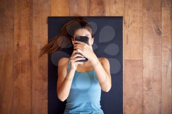 Overhead View Of Woman Lying On Exercise Mat Wearing Wireless Earphones Connected To Mobile Phone