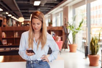 Casually Dressed Young Businesswoman Checking Mobile Phone In Modern Open Plan Workplace