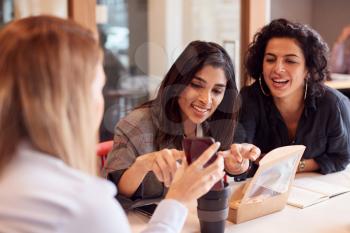 Group Of Young Businesswomen Sitting Around Table Having Working Lunch And Looking At Mobile Phones