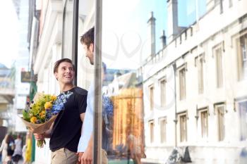 Loving Male Gay Couple Holding Hands Coming Out Of Florists Holding Bunch Of Flowers