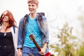 Excited Young Couple Carrying Camping Equipment And Guitar Through Field To Music Festival