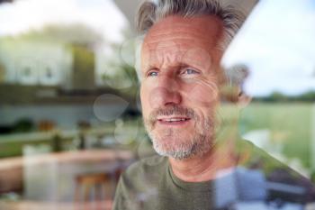 Senior Man Standing And Looking Out Of Kitchen Door Viewed Through Window