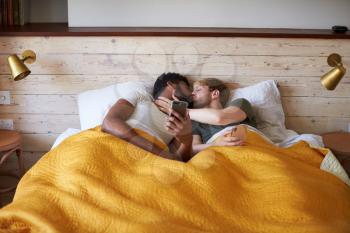 Male Gay Couple Lying In Bed At Home Checking Mobile Phones Together