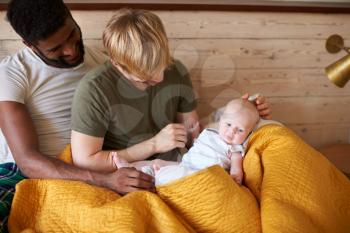 Loving Male Same Sex Couple Cuddling Baby Daughter In Bedroom At Home Together