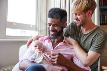 Loving Male Same Sex Couple Cuddling Baby Daughter On Sofa At Home Together