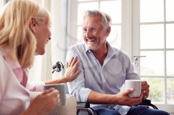 Mature Couple With Man In Wheelchair Sitting In Lounge At Home Talking Together