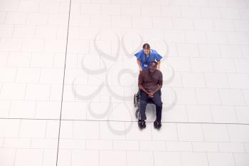 Overhead View Of Nurse Wheeling Male Patient In Wheelchair Through Lobby Of Modern Hospital Building
