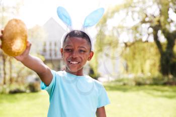 Portrait Of Boy Wearing Bunny Ears Holding Chocolate Egg On Easter Egg Hunt In Garden