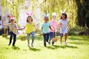 Group Of Children Wearing Bunny Ears Running To Pick Up Chocolate Egg On Easter Egg Hunt In Garden