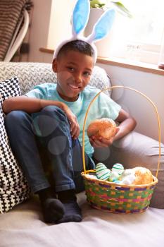 Boy Wearing Bunny Ears Sitting On Sofa Eating Chocolate Egg He Has Found On Easter Egg Hunt
