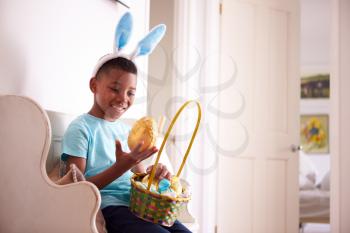 Boy Wearing Bunny Ears Sitting On Seat Holding Chocolate Egg He Has Found On Easter Egg Hunt