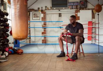 Male Boxer Training In Gym Sitting Next To Boxing Ring Putting Wraps On Hands