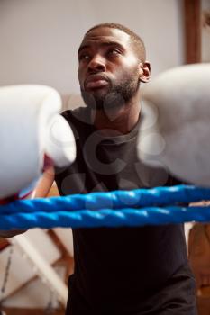 Male Boxer In Gym Wearing Boxing Gloves Leaning On Ropes Of Boxing Ring