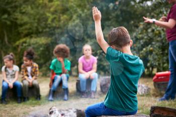 Children On Outdoor Activity Camping Trip Sit Around Camp Fire With Arms Raised Answering Question
