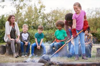 Female Team Leader With Group Of Children On Outdoor Activity Trip Cooking Meal Over Camp Fire