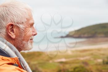 Profile Shot Of Active Senior Man Walking Along Coastal Path In Winter With Beach And Cliffs Behind