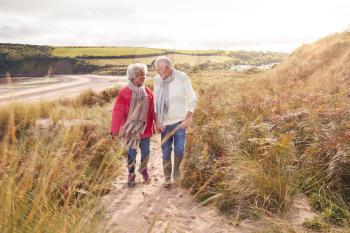 Loving Active Senior Couple Walking Arm In Arm Through Sand Dunes On Winter Beach Vacation