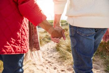 Close Up Of Loving Senior Couple Holding Hands Walking Through Sand Dunes On Winter Vacation