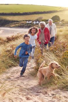 Grandchildren And Pet Dog Exploring Sand Dunes With Grandparents On Winter Beach Vacation