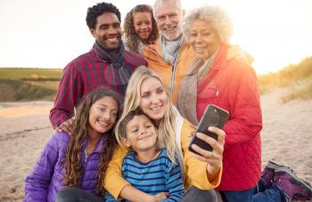 Multi-Generation Family Sitting By Fire On Winter Beach Vacation Taking Selfie