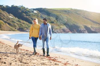 Loving Couple Holding Hands As They Walk With Dog Along Shoreline On Winter Beach Vacation