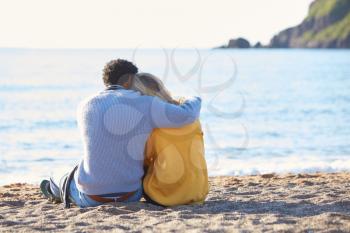 Rear View Of Loving Couple Hugging As They Sit On Sand By Shoreline On Winter Beach Vacation