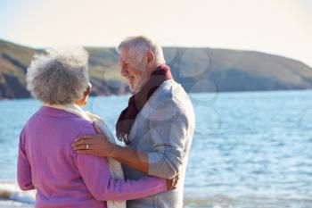 Loving Retired Couple Hugging Standing By Shore On Winter Beach Vacation Against Flaring Sun