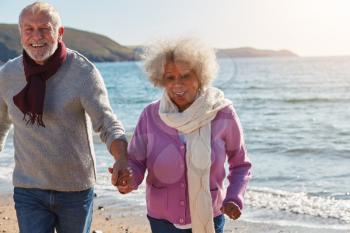 Active Senior Couple Running Along Winter Beach By Waves Holding Hands