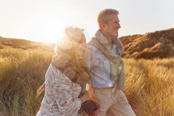 Loving Retired Couple Walking Arm In Arm Through Sand Dunes On Beach Vacation Against Flaring Sun