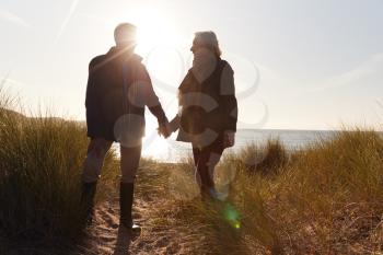 Silhouette Of Loving Senior Couple Holding Hands As They Walk Along Coast Path Against Flaring Sun