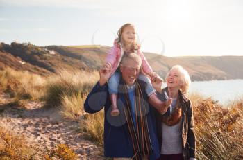 Grandfather Giving Granddaughter Ride On Shoulders As They Walk Through Sand Dunes With Grandmother