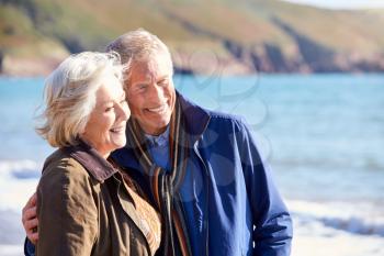 Loving Senior Couple Hugging As They Walk Along Shoreline Of Beach By Waves