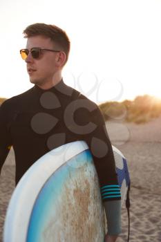 Young Man In Sunglasses Wearing Wetsuit Enjoying Surfing Staycation On Beach As Sun Sets