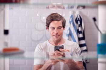 View Through Bathroom Cabinet Of Man Checking Messages On Phone Before Going To Work