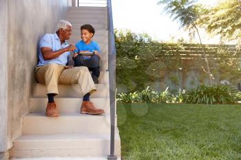 Grandfather With Grandson Sitting On Steps Outdoors At Home Using Digital Tablet