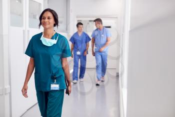 Female Doctor Wearing Scrubs In Hospital Walking Along Corridor Holding Digital Tablet