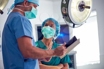 Male And Female Surgeons Wearing Scrubs Looking At Digital Tablet In Hospital Operating Theater