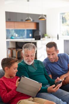 Multi-Generation Male Hispanic Family Sitting On Sofa At Home Using Mobile Phones And Digital Tablet