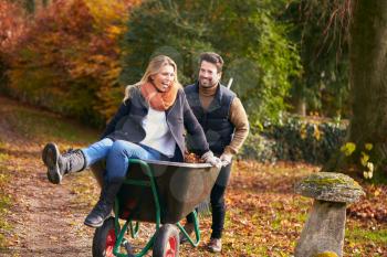 Man Pushing Woman In Wheelbarrow As Couple Rake Autumn Leaves From Garden Together