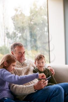 Grandchildren On Sofa At Home Showing Grandfather How To Use Mobile Phone