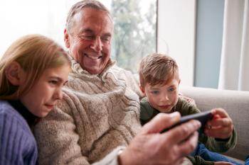 Grandchildren On Sofa At Home Showing Grandfather How To Use Mobile Phone