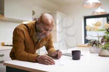Mature Man Reviewing And Signing Domestic Finances And Investment Paperwork In Kitchen At Home