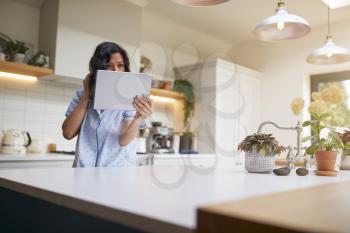 Mature Hispanic Woman In Pyjamas At Home In Kitchen Checks Hair And Appearance With Digital Tablet