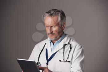 Studio Portrait Of Mature Male Doctor In White Coat Using Digital Tablet Against Grey Background