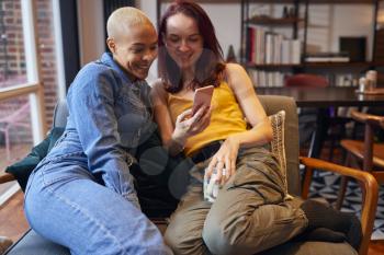Loving Same Sex Female Couple Sitting On Sofa At Home Looking At Mobile Phone Together