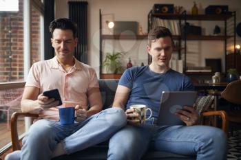 Same Sex Male Couple Sitting On Sofa Using Digital Tablet And Mobile Phone As They Relax At Home