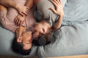 Overhead Shot Of Loving Same Sex Male Couple Lying On Bed At Home Hugging And Kissing Together